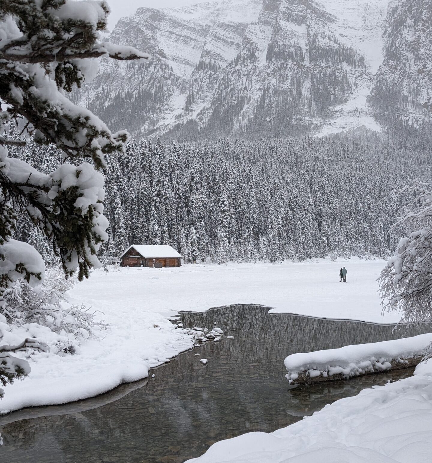 Couple walking hand in hand across frozen Lake Louise in winter, with the Lake Louise boathouse and icy stream visible in Banff National Park