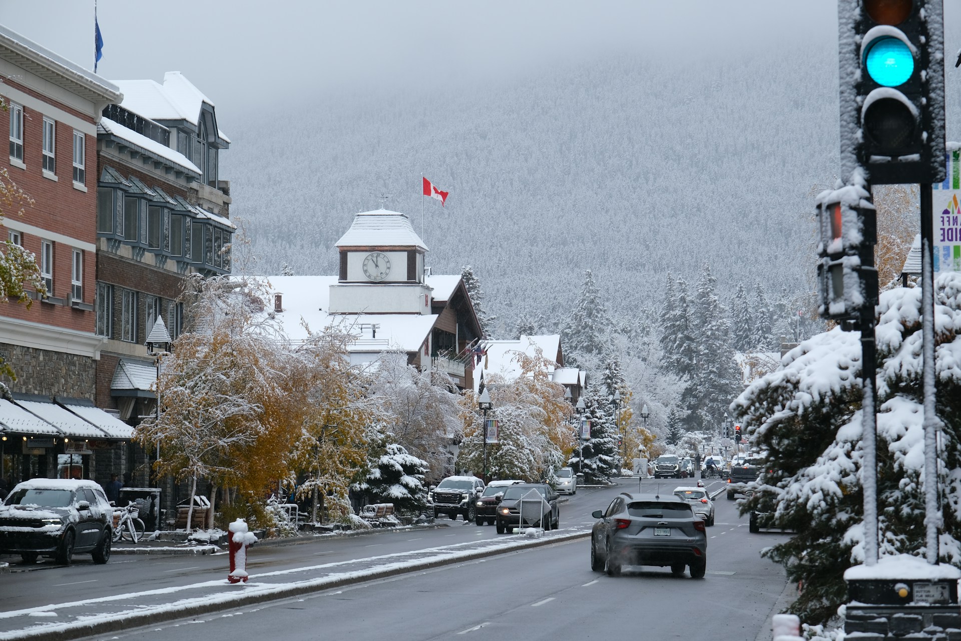 Fresh snow on Banff Avenue with golden autumn leaves still on the trees, shopfronts, parked cars and a fog-shrouded forested slope behind