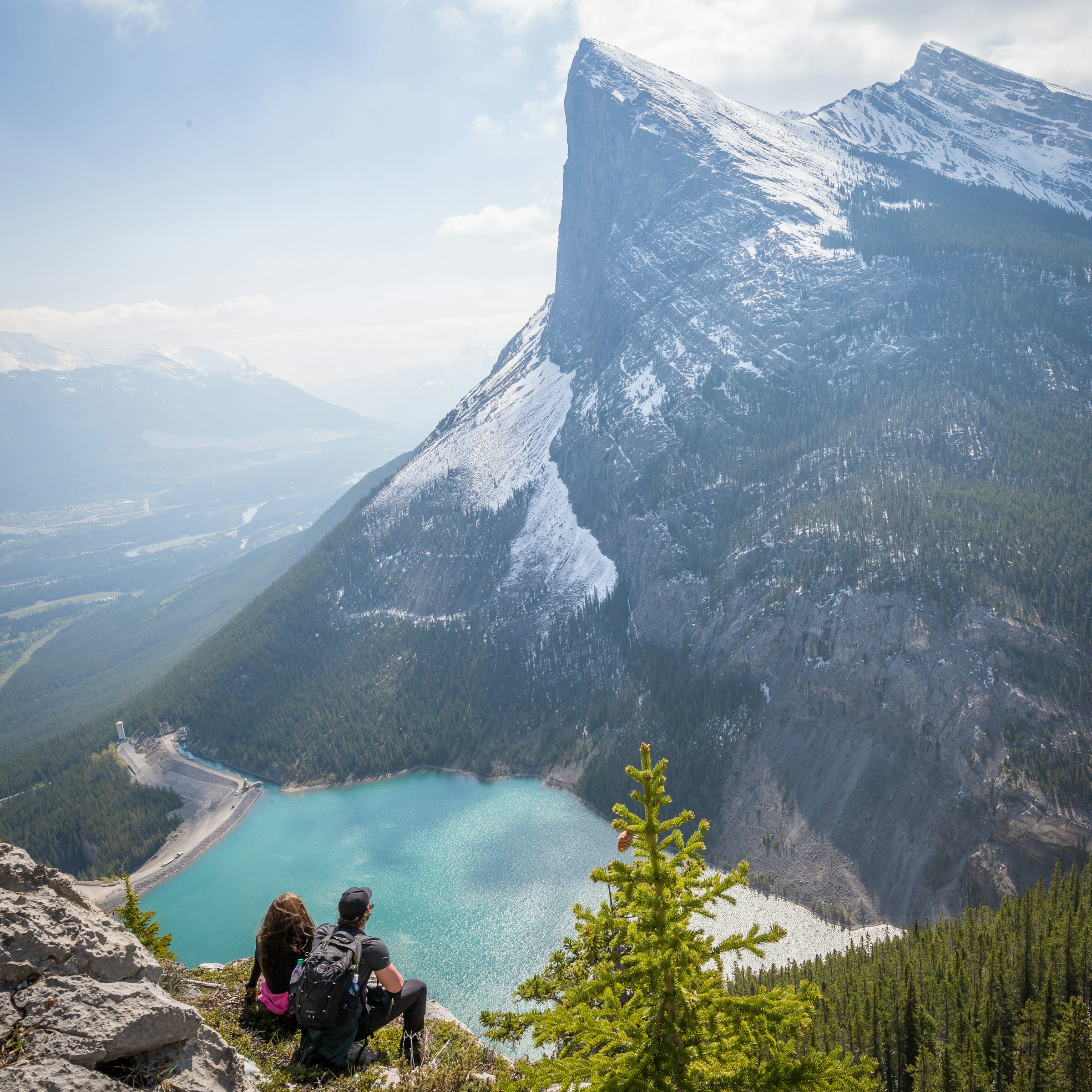 Two hikers sitting on a rocky ledge overlooking a turquoise reservoir and a steep peak near Canmore, Alberta