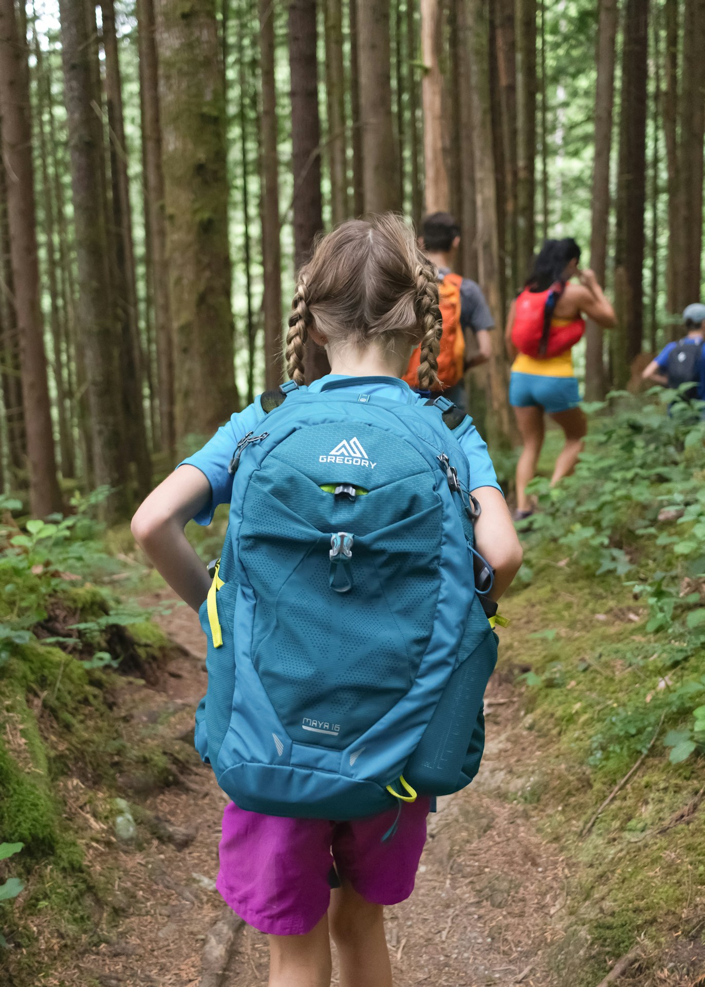 A young girl in braids carrying her own blue day-pack along a forest trail, with adults walking ahead of her