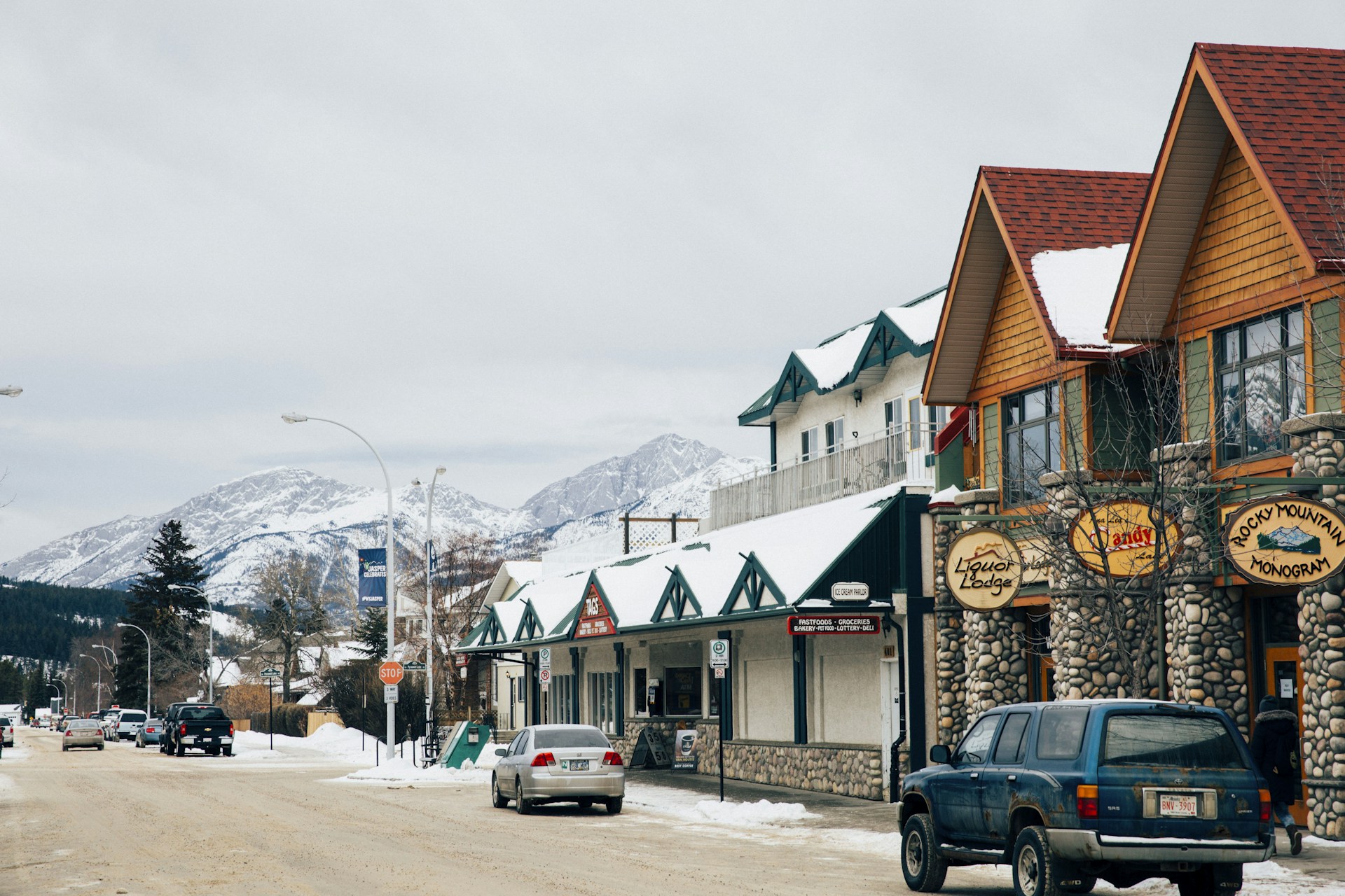 A snowy downtown Jasper street with timber-and-stone storefronts, parked cars and a snow-covered mountain behind