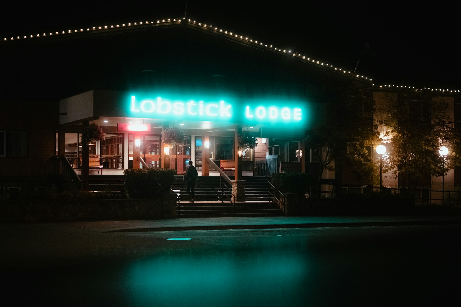 Lobstick Lodge in Jasper at night, its teal neon sign lit above the entrance steps and string lights along the roofline