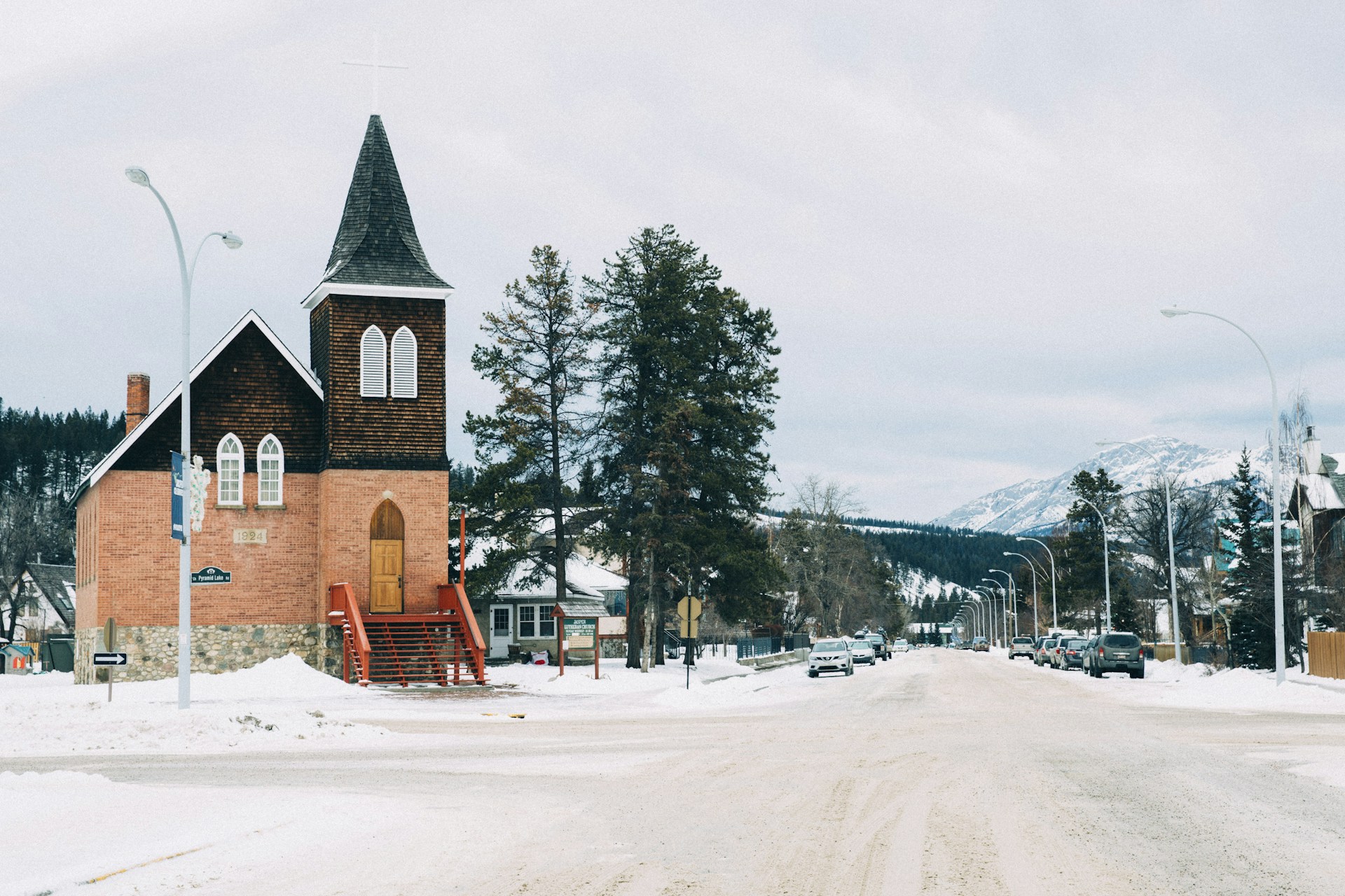 A red-brick church with a shingled steeple on a snowy corner in Jasper townsite, a wide street leading toward snow-dusted mountains