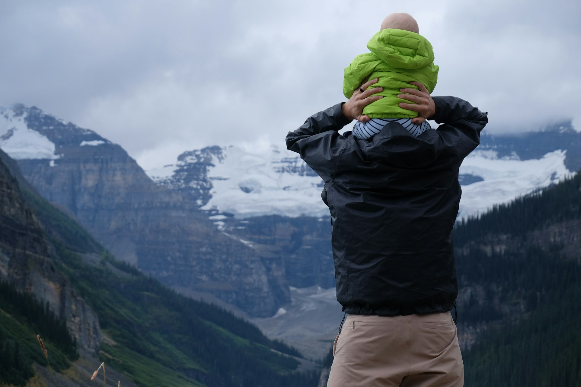 A parent in a rain jacket holding a toddler on his shoulders, looking across a forested valley to the Victoria Glacier above Lake Louise