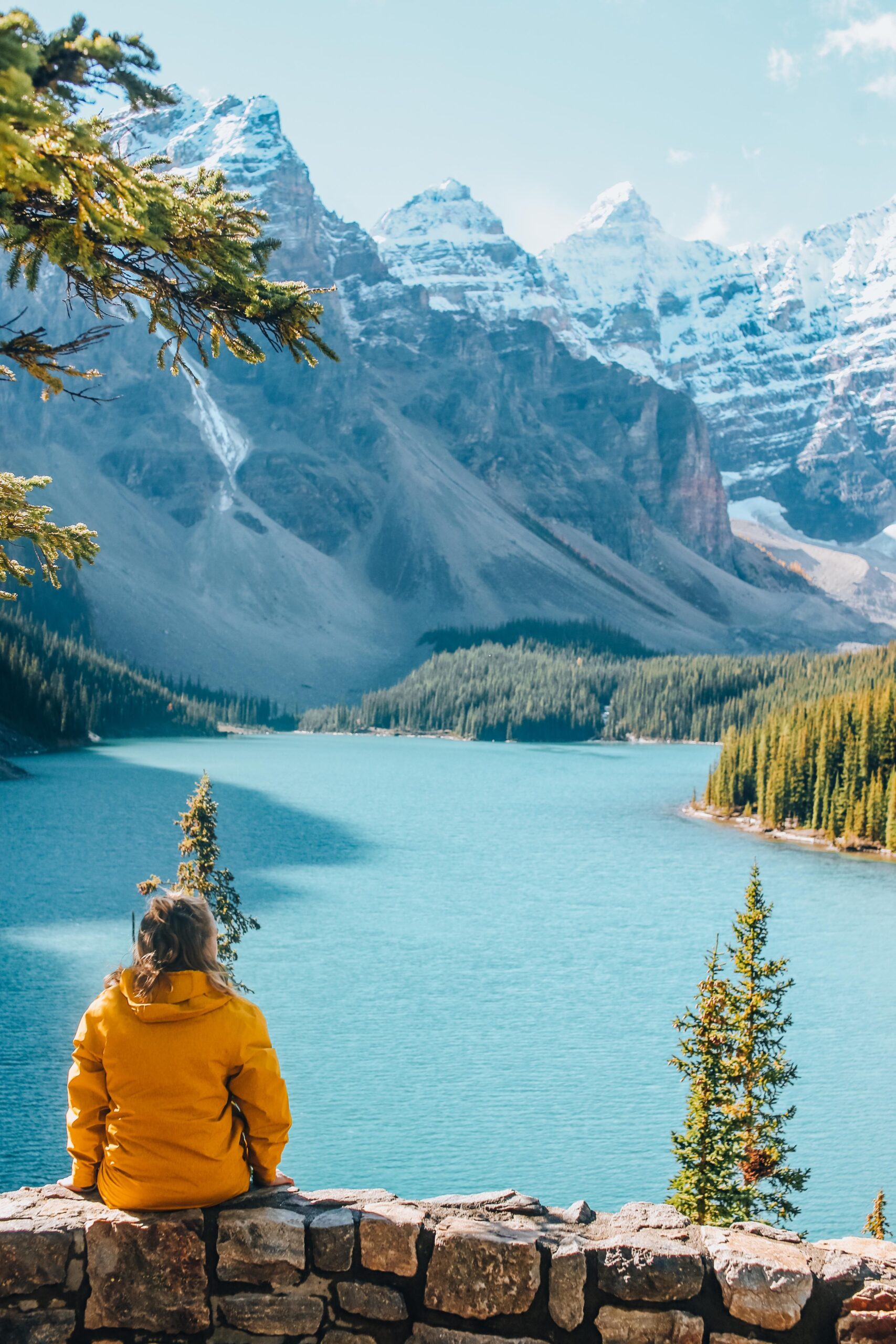 A visitor in a yellow jacket sitting on the Rockpile wall above Moraine Lake, looking out over turquoise water toward the snow-dusted Valley of the Ten Peaks