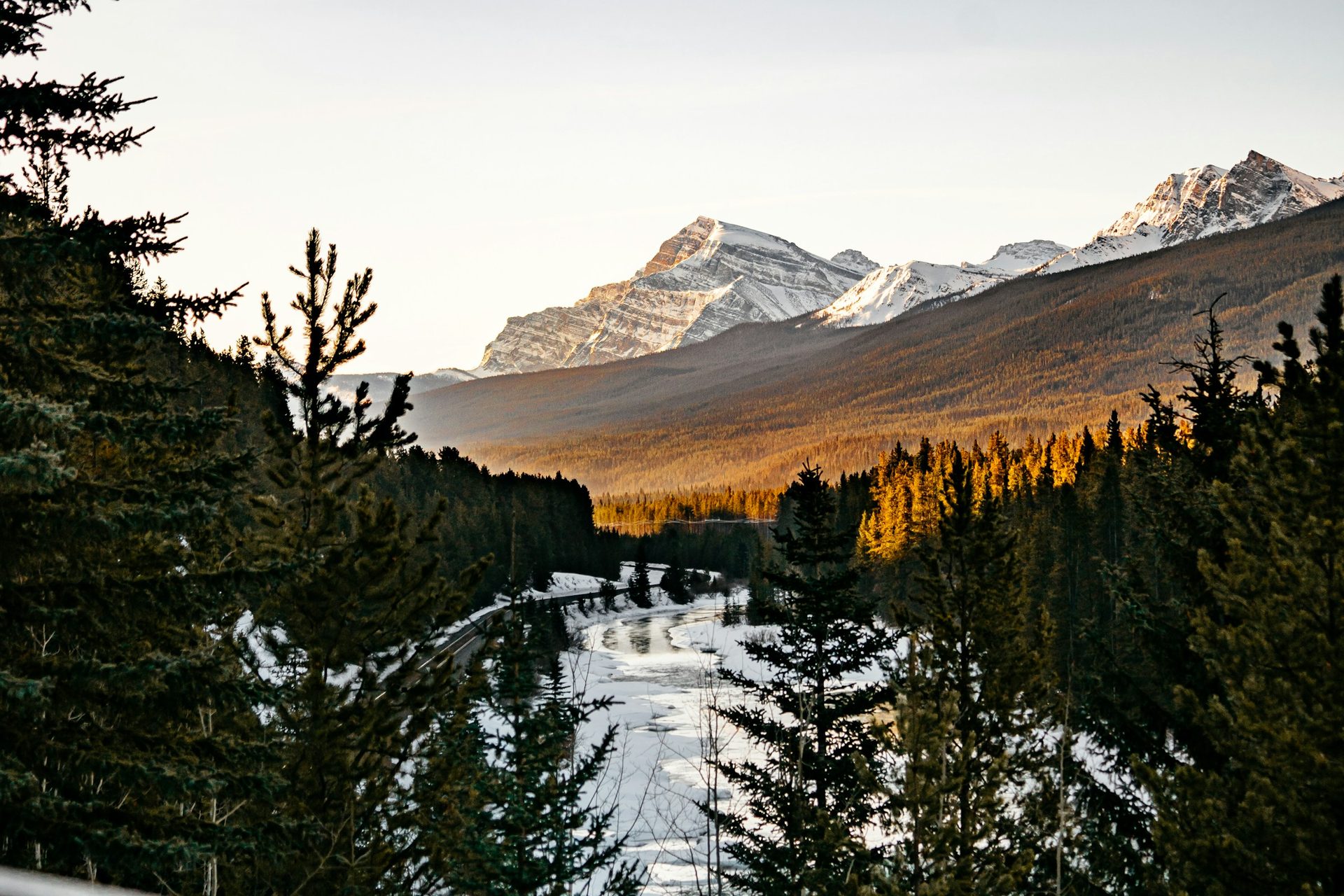 Low sun lighting snow-capped peaks above a snow-covered, partly frozen river winding through dark spruce forest near Banff