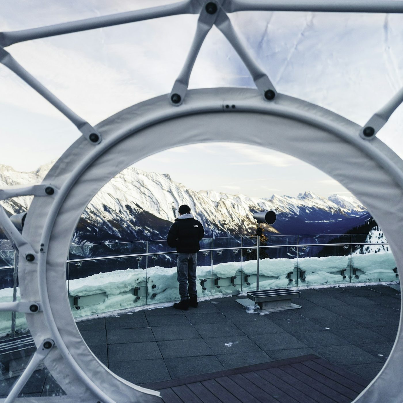 A visitor in a winter jacket at the rail of the Banff Gondola rooftop deck, framed by a large white viewing ring, with snowy peaks beyond