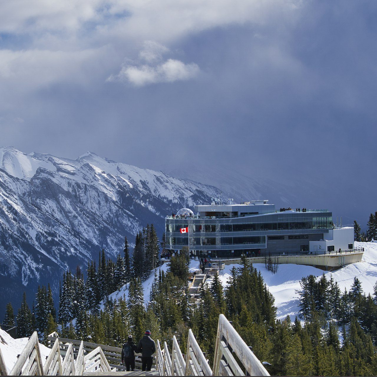 Visitors on the snowy Sulphur Mountain boardwalk stairs below the Banff Gondola summit building, with snow-covered peaks under a cloudy sky