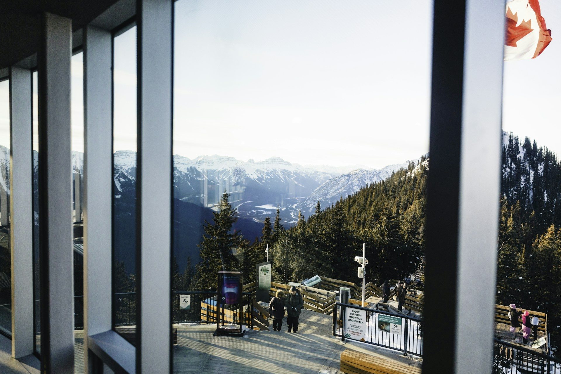 View through the tall windows of the Banff Gondola summit building to the outdoor deck, visitors and snowy mountain ranges