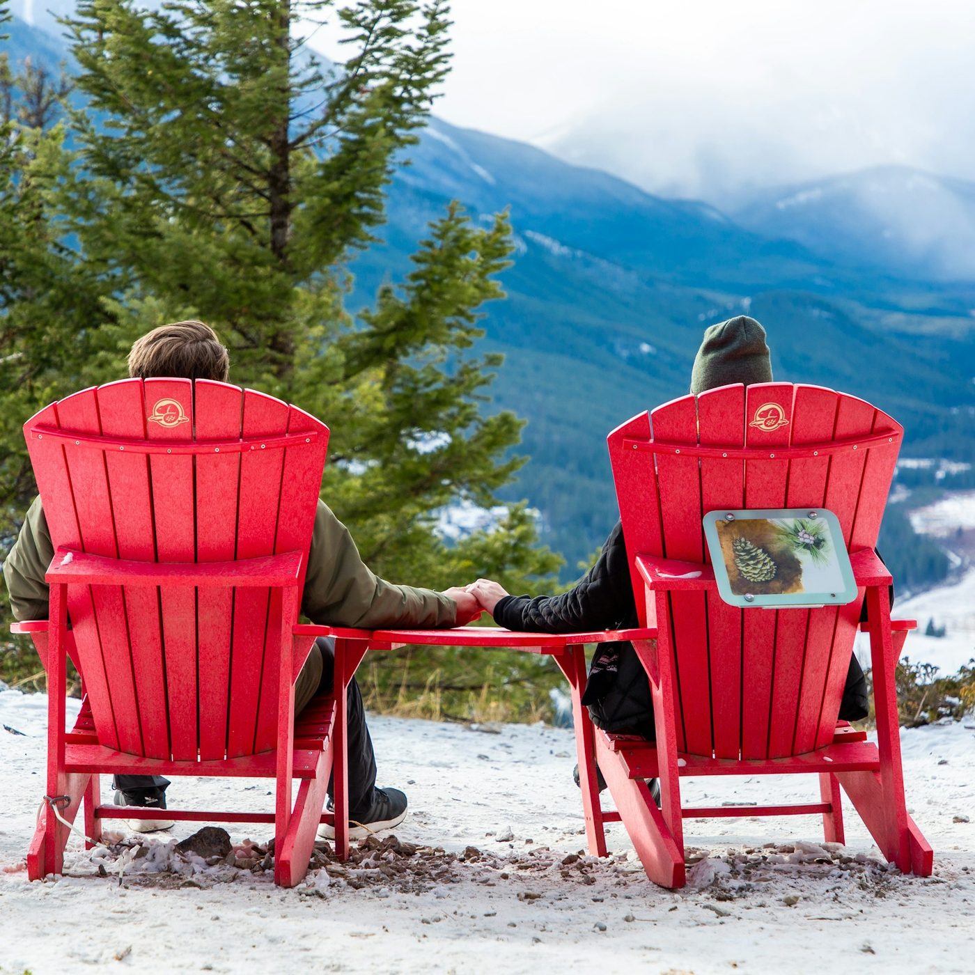 Two people sitting in Parks Canada red chairs on a snowy viewpoint, looking down the Bow Valley toward snow-covered mountains