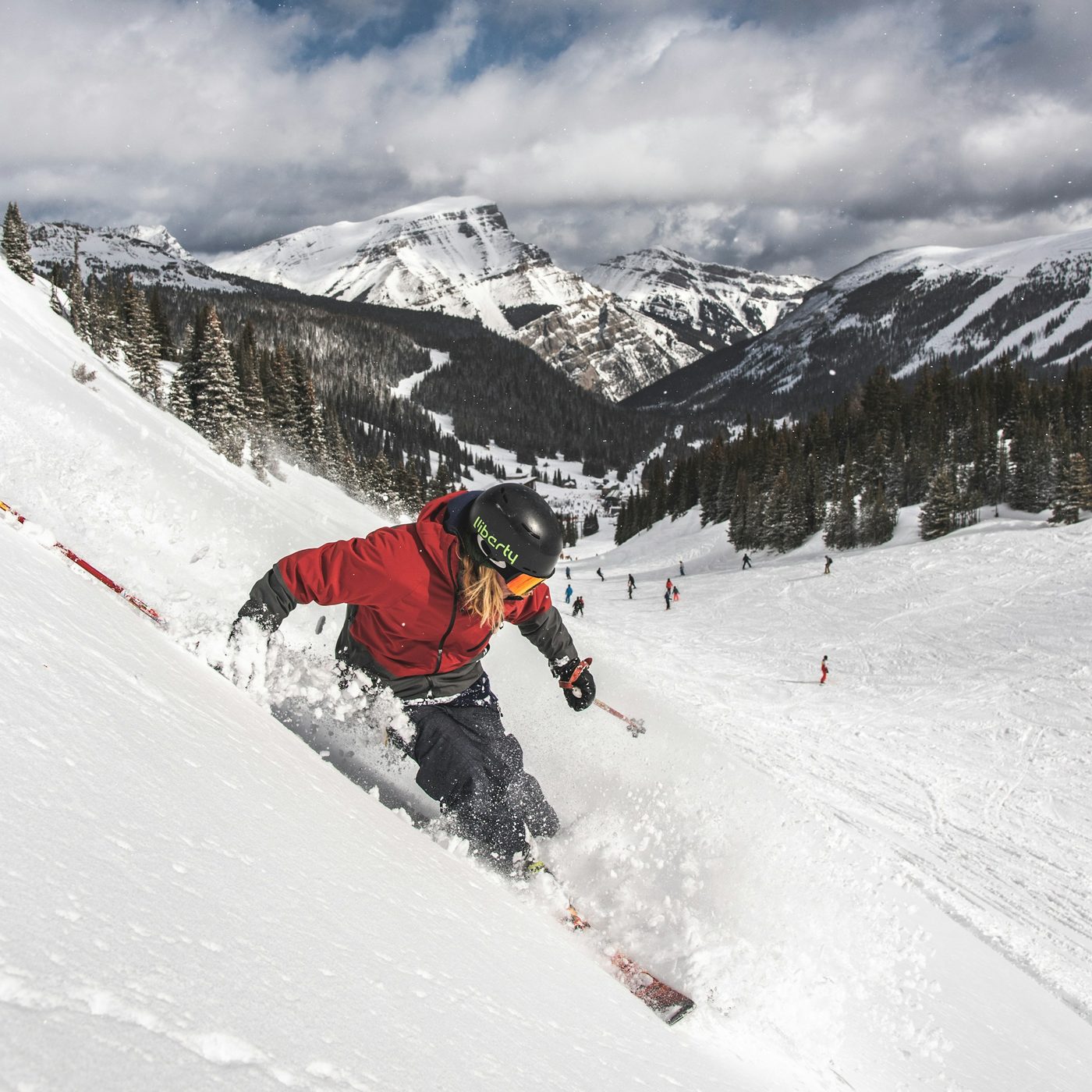 A skier in a red jacket turning through deep powder on a steep slope at Banff Sunshine Village, with skiers on the run below and snowy peaks behind