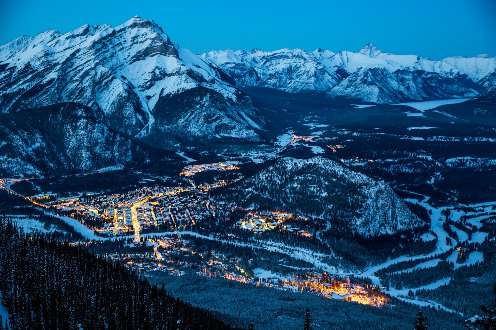 Banff townsite lit up at dusk in winter, seen from above, with snow-covered peaks around the Bow Valley
