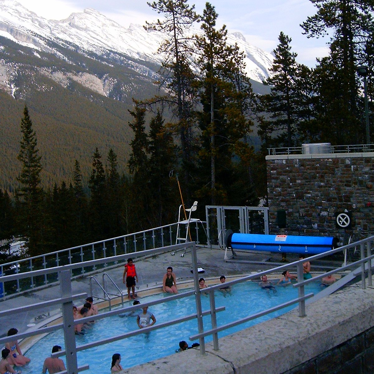 People soaking in the outdoor pool at the Banff Upper Hot Springs, with forest and snowy peaks beyond the railing