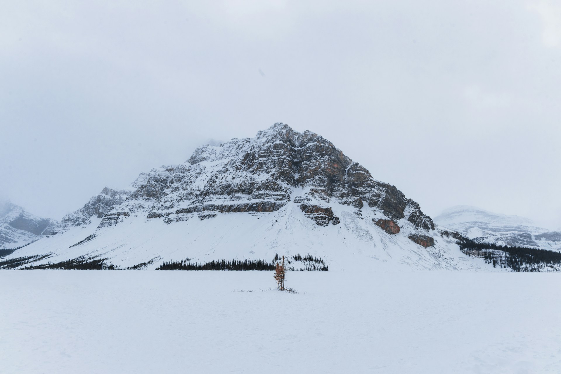 A lone person standing on snow-covered, frozen Bow Lake below a snowy pyramid-shaped peak under grey cloud
