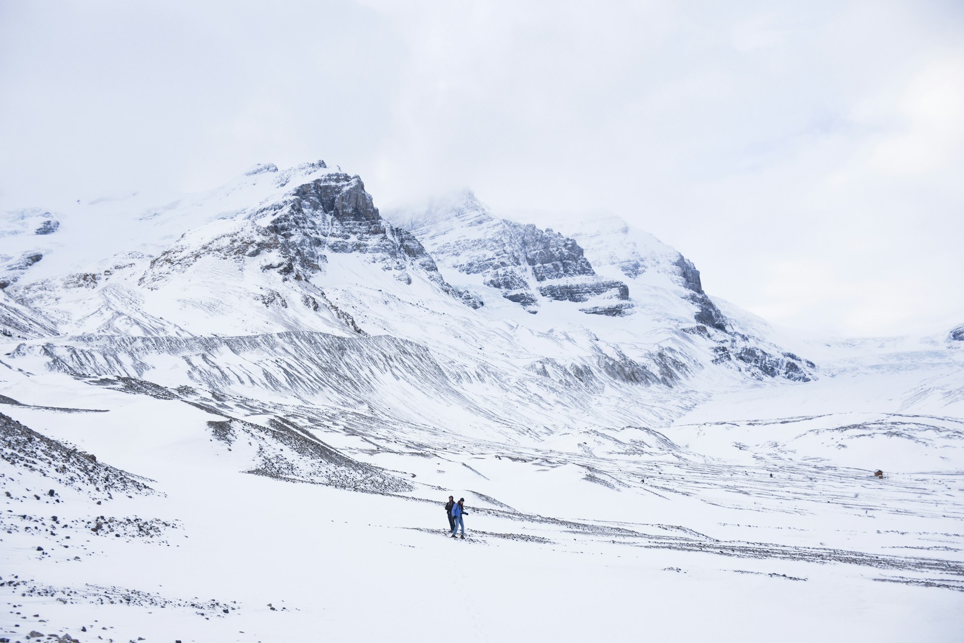 Two walkers crossing deep snow below the snow-covered moraines and peaks of the Columbia Icefield under low winter cloud
