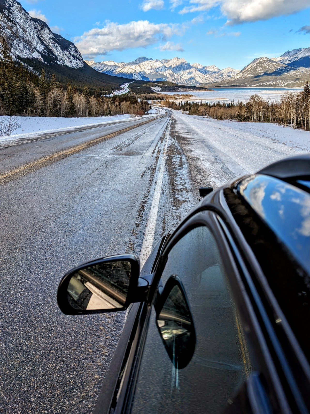 View past a car's side mirror down a snow-edged highway toward frozen Abraham Lake and snow-covered peaks in December