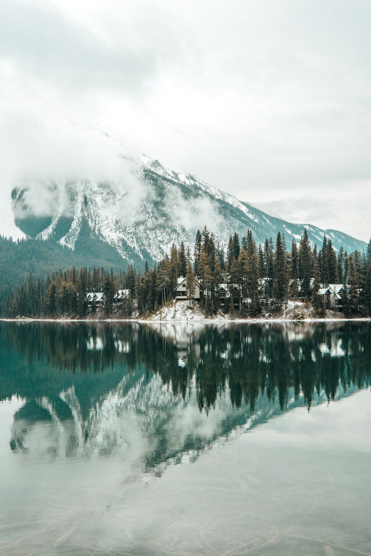 Emerald Lake Lodge cabins dusted with early snow on a forested point, mirrored in still water below a cloud-wrapped peak