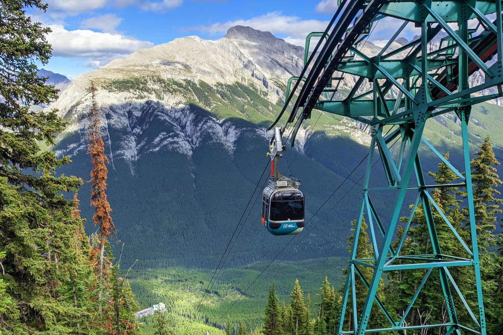 Banff Gondola cabin climbing past the upper-terminal tower on Sulphur Mountain, with forested slopes and a rocky peak behind