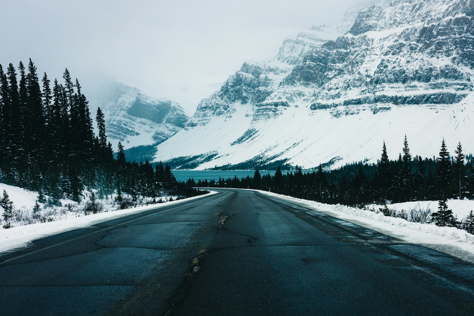 A cleared two-lane road running between snowbanks and dark spruce forest toward snow-covered peaks in Banff National Park