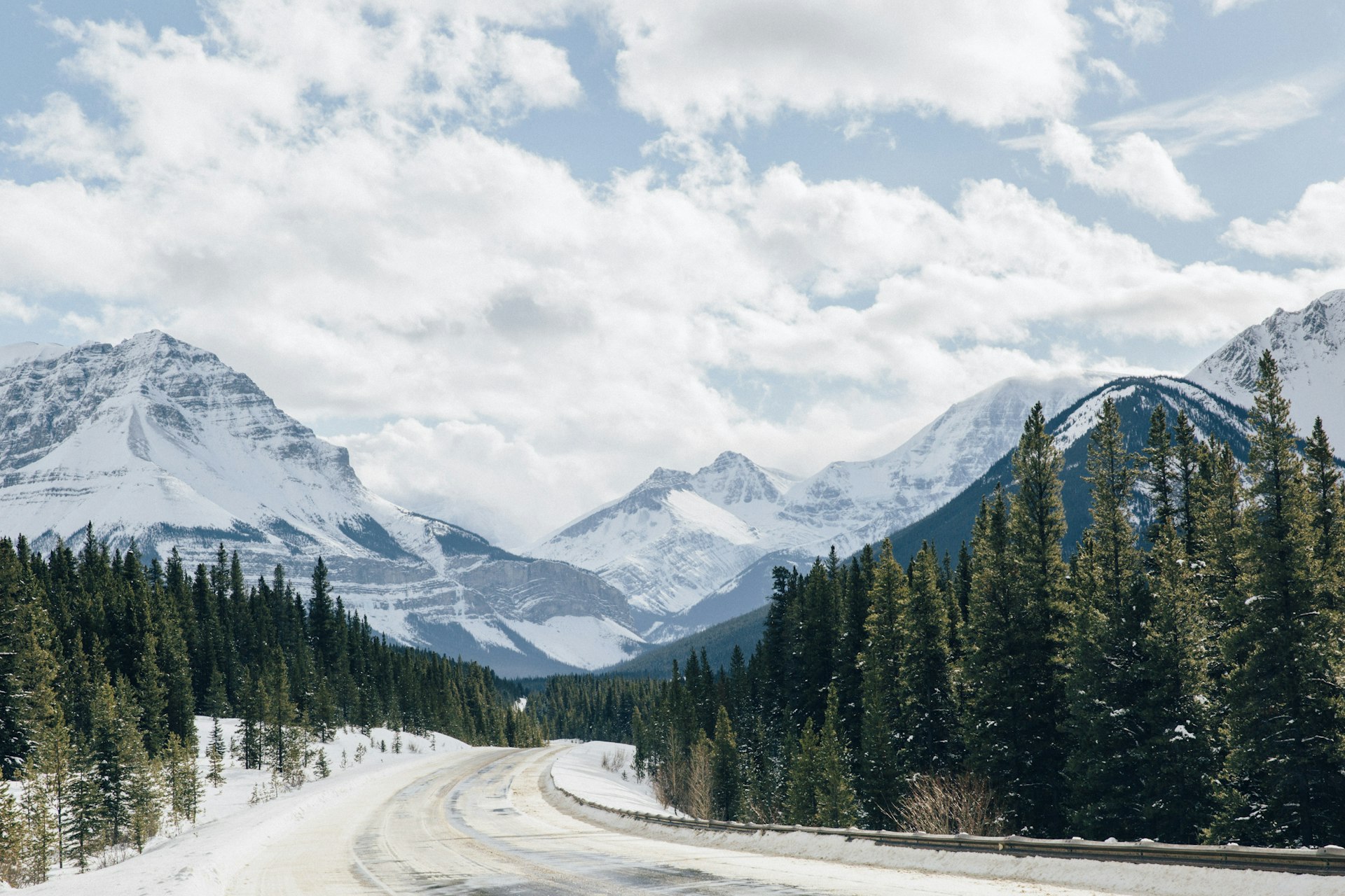 The Icefields Parkway in winter, a snow-packed road curving between spruce forest toward snow-covered peaks under scattered cloud