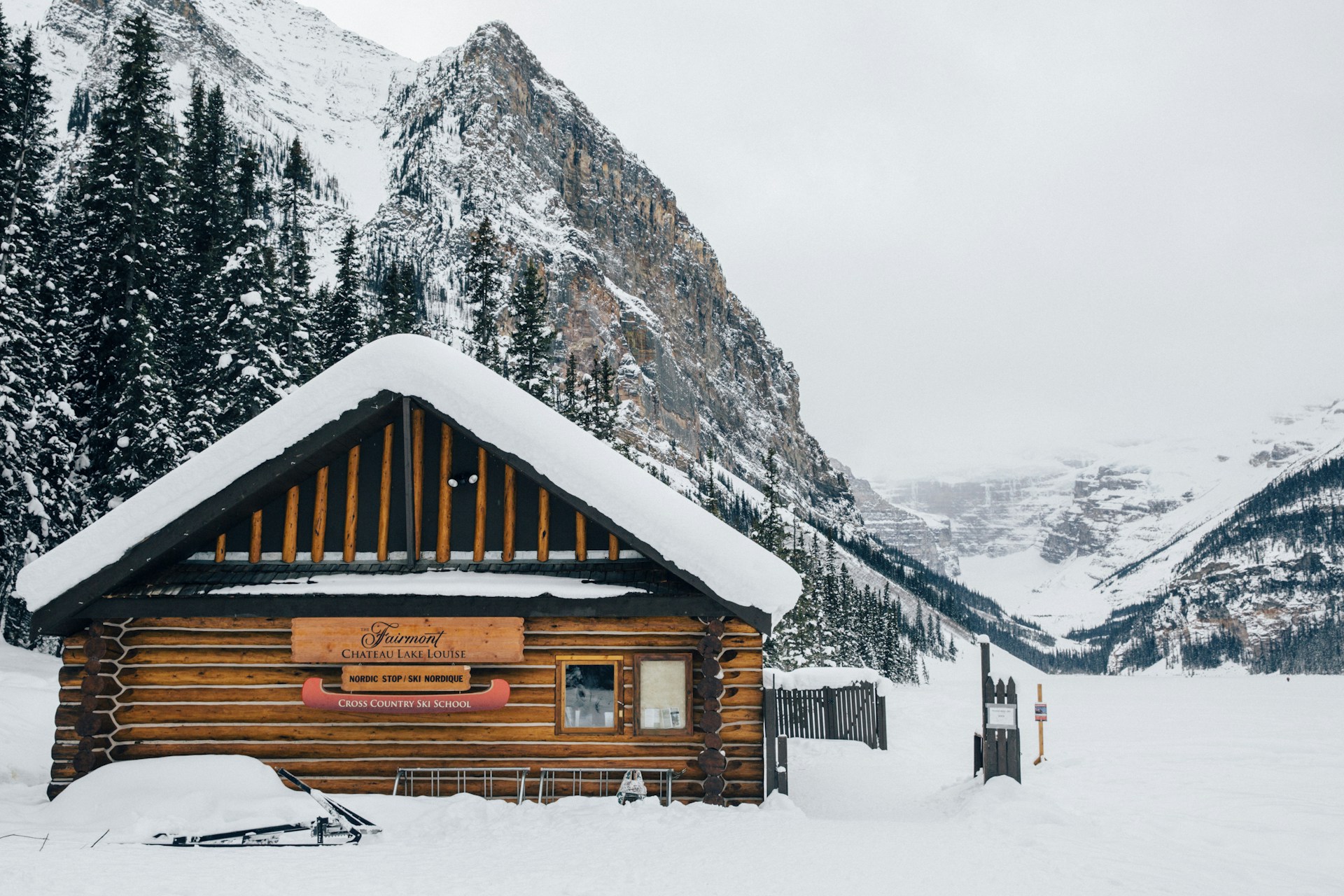 The snow-covered log cabin of the Lake Louise cross-country ski school on the lakeshore, with the frozen lake and steep peaks behind