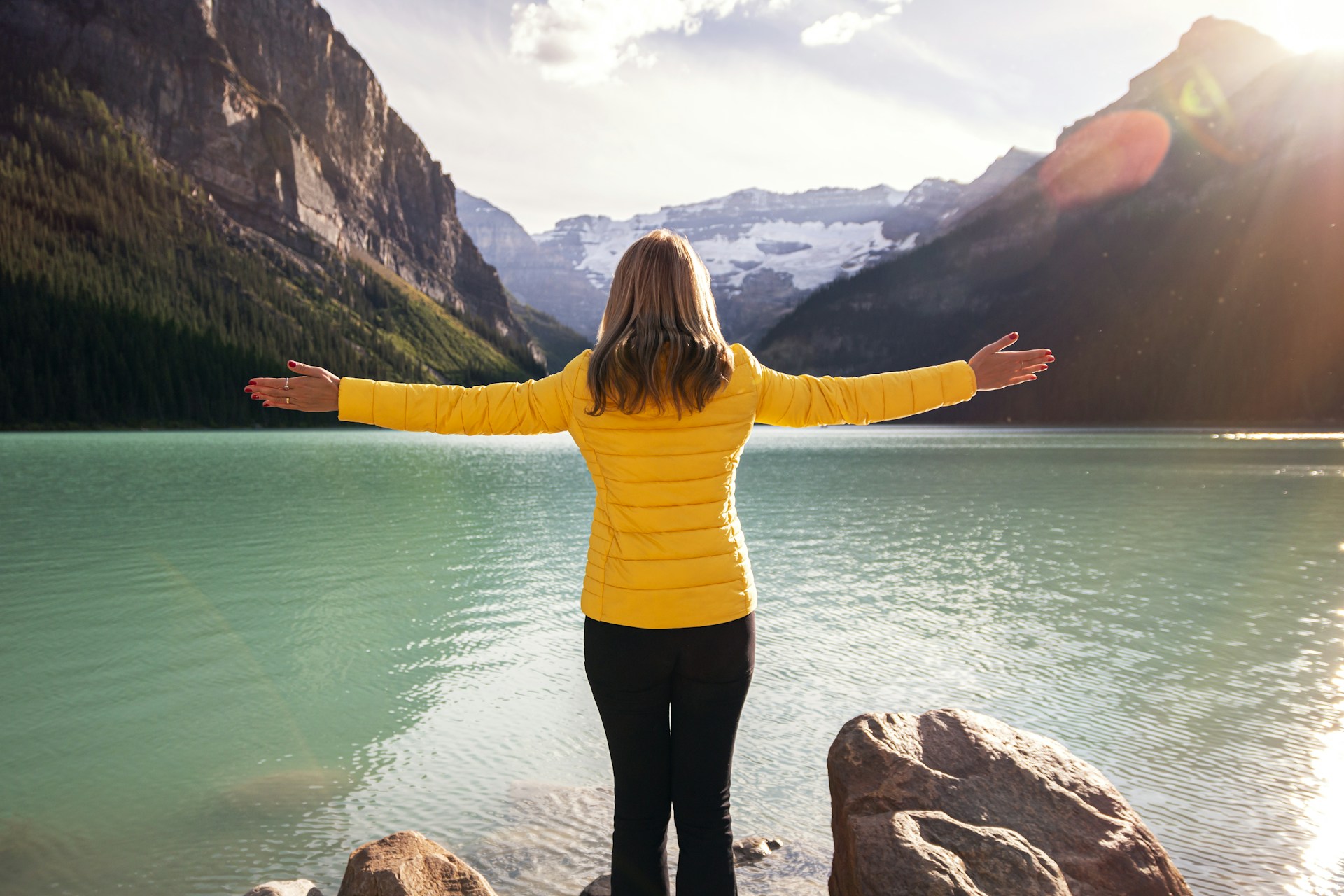 A woman in a yellow jacket standing on the lakeshore rocks at Lake Louise, arms outstretched toward Victoria Glacier