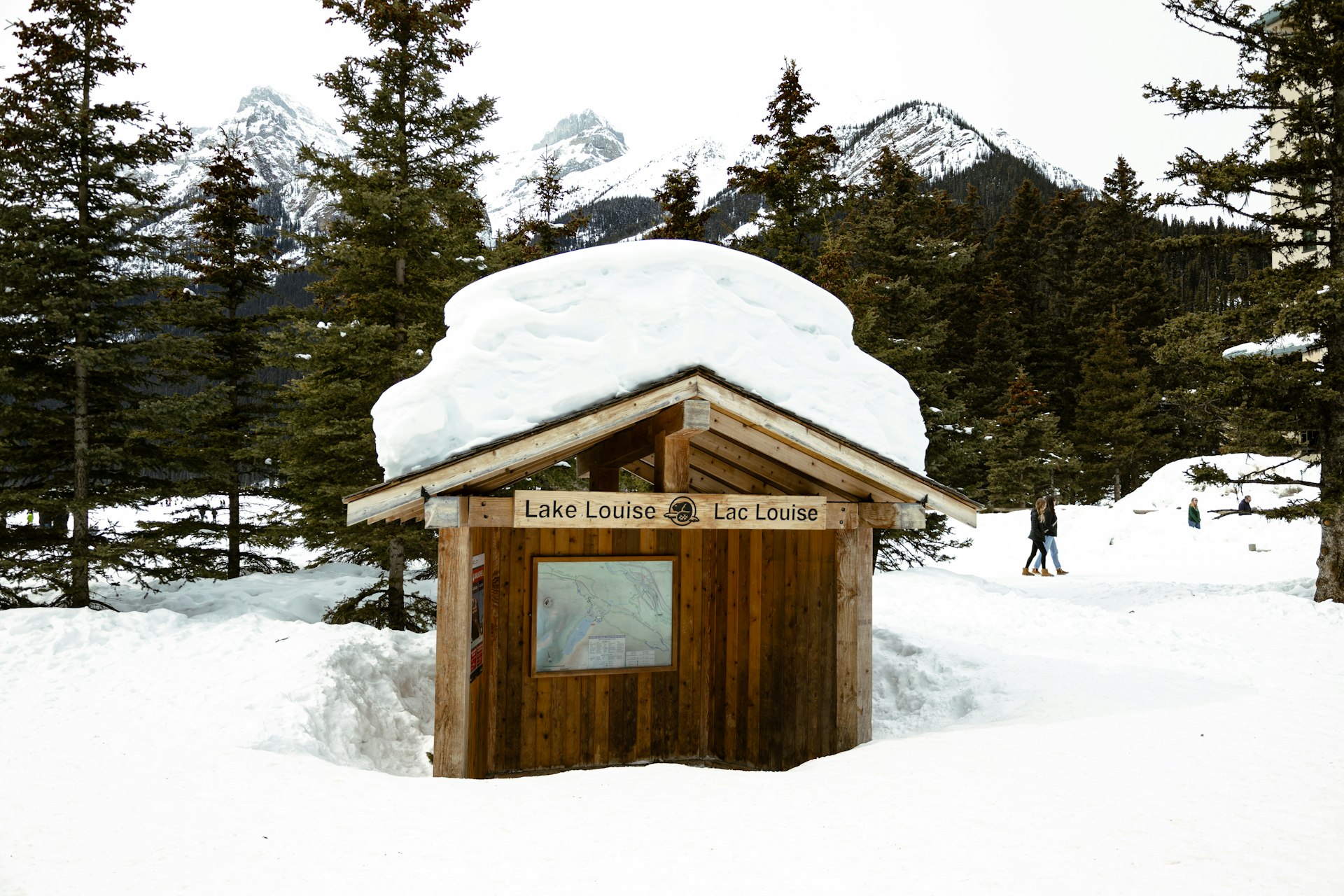 A wooden Lake Louise trail kiosk with a deep cap of snow on its roof, walkers on the packed trail behind and spruce-covered mountains beyond