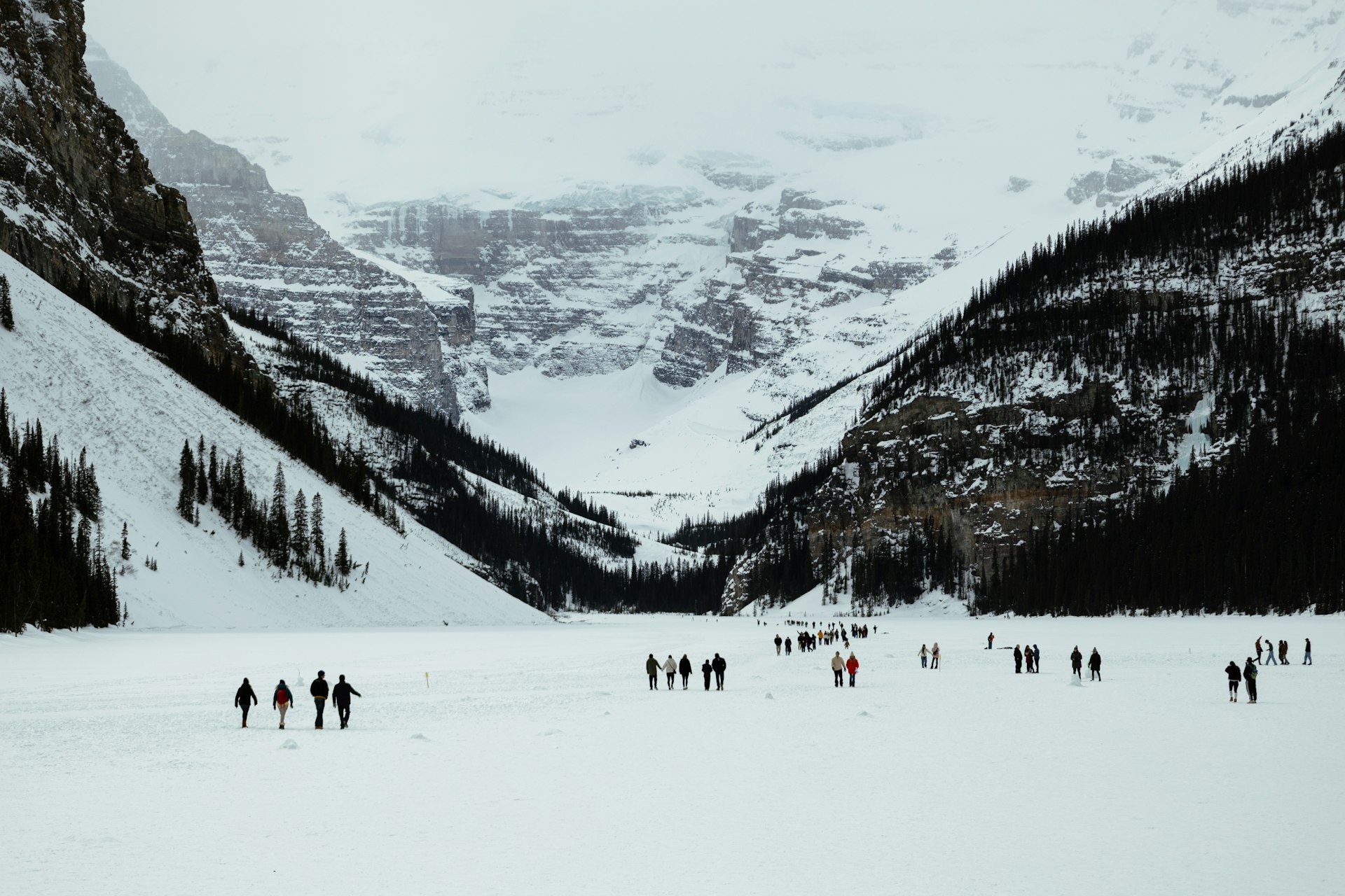 Walkers strung out across snow-covered, frozen Lake Louise, heading toward Victoria Glacier between steep forested slopes