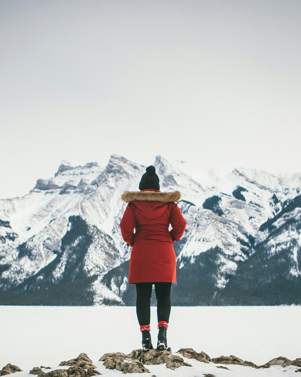 A person in a red parka stands on a rock above frozen, snow-covered Lake Minnewanka, facing snowy peaks