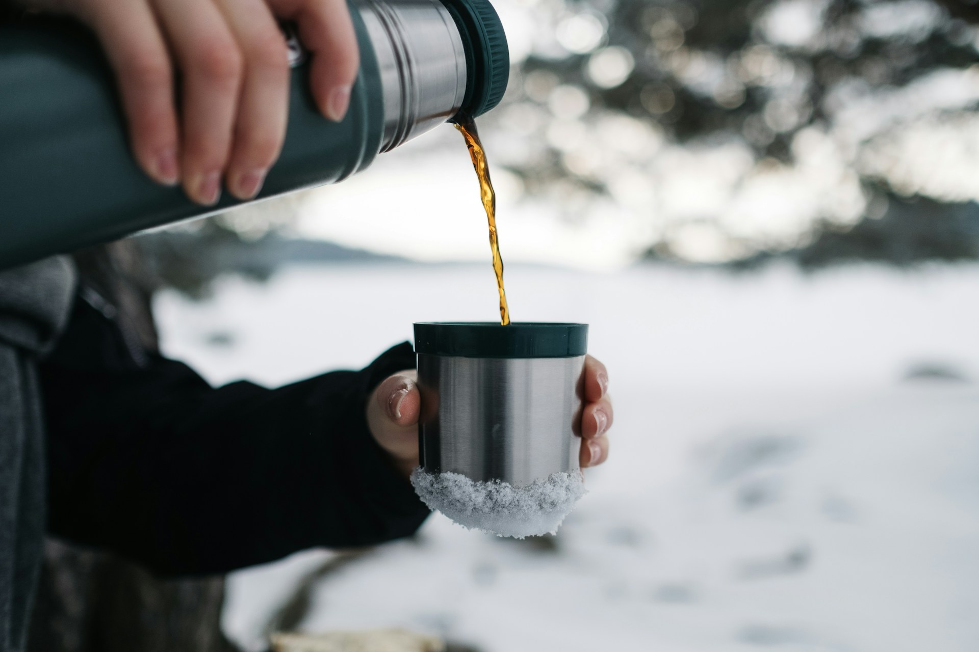 A gloved hand pouring a hot drink from a steel flask into its cup in the snow