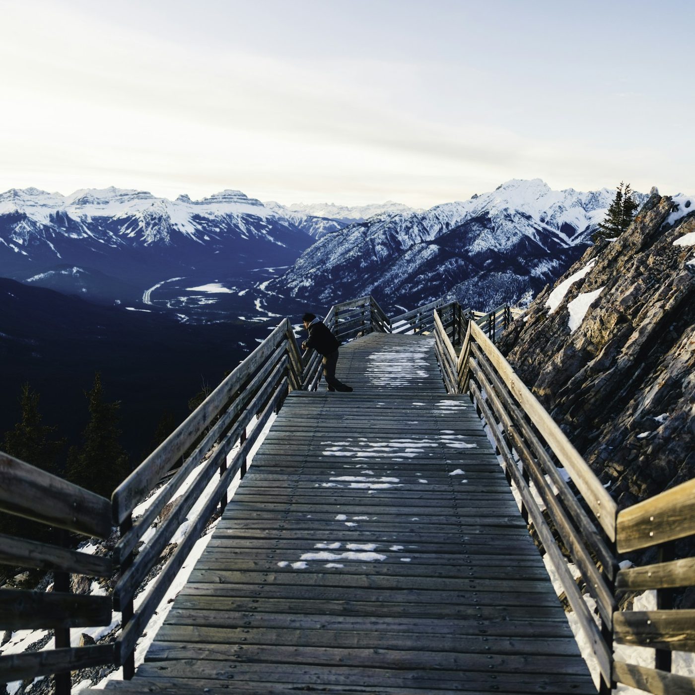 Wooden boardwalk steps running down a snowy ridge on Sulphur Mountain toward a range of snow-covered peaks
