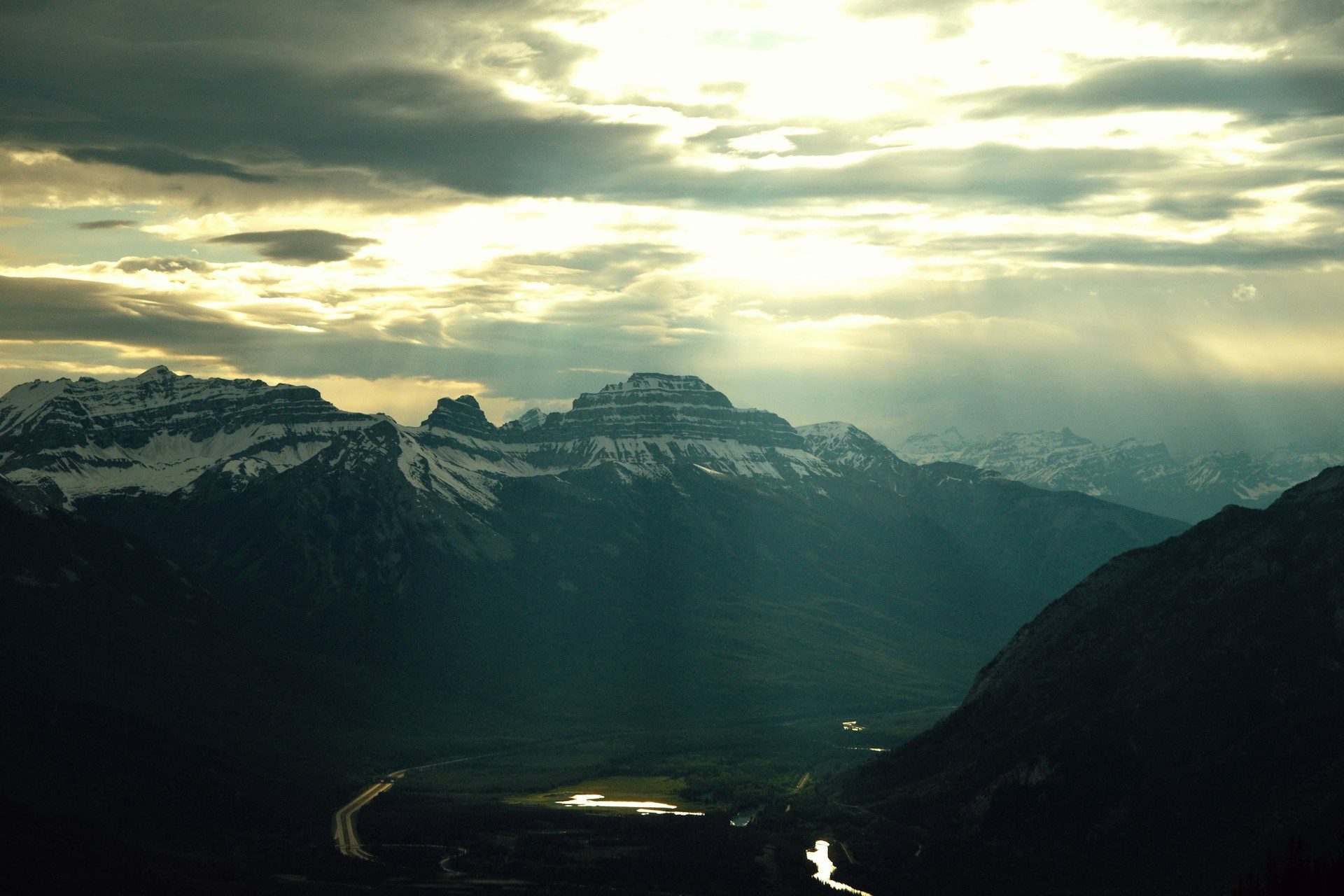 Late-day sun breaking through clouds over dark mountain ridges and a valley, seen from the top of Sulphur Mountain