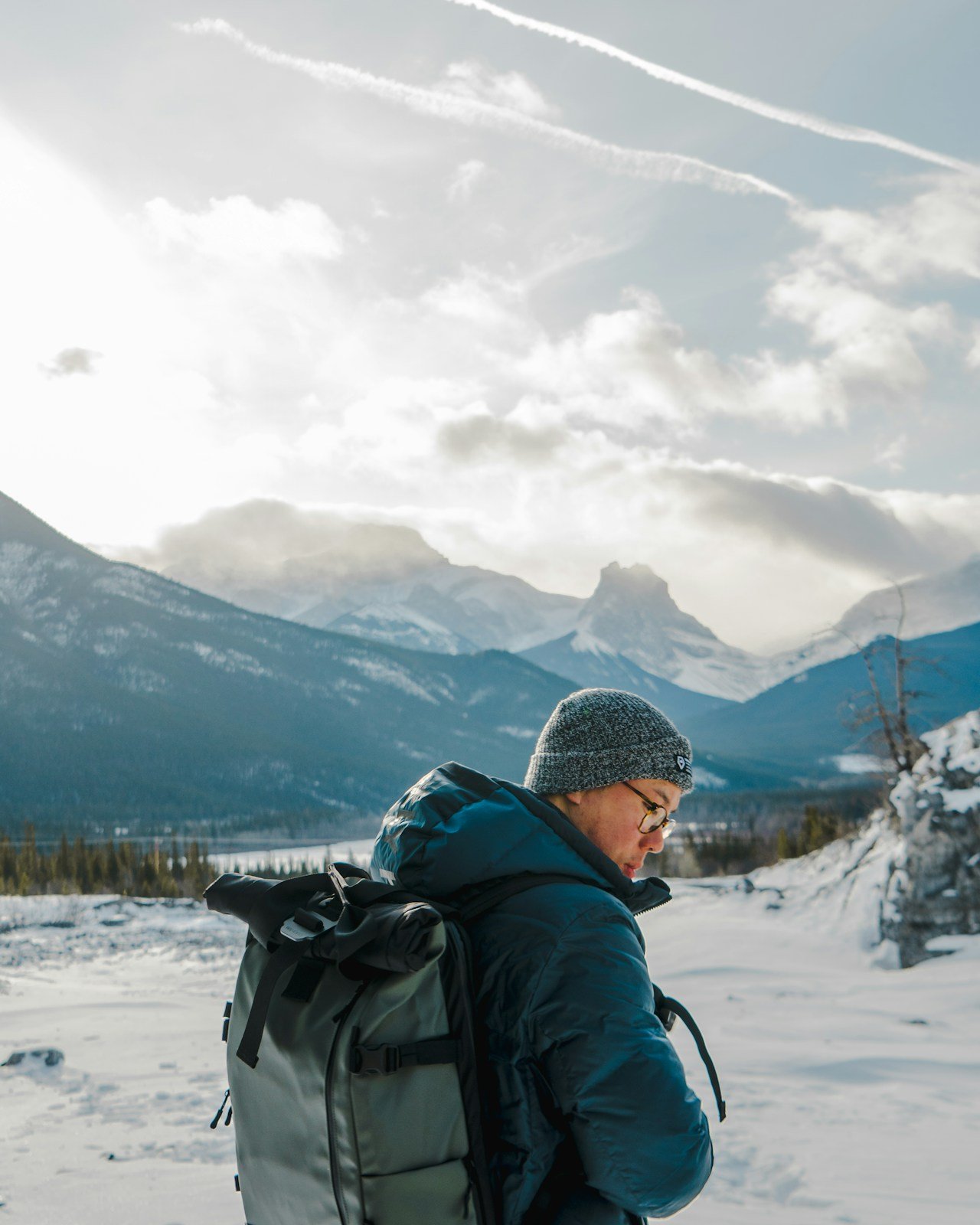 A traveller in a grey toque, glasses and a heavy parka with a backpack, standing in snow below cloud-wrapped peaks near Banff